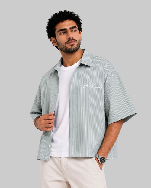 A man wearing a light sage green and white vertically striped short-sleeve button-up shirt over a white t-shirt, paired with beige pants, against a plain white background.