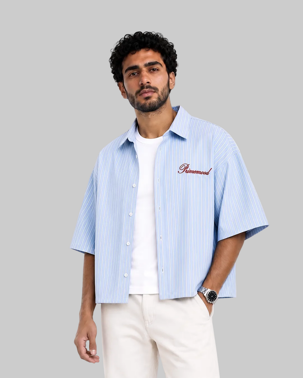 A man wearing a light blue and white vertically striped short-sleeve button-up shirt with '' embroidered in red on the chest, layered over a white t-shirt and cream pants, against a plain white background. boxy shirt blue