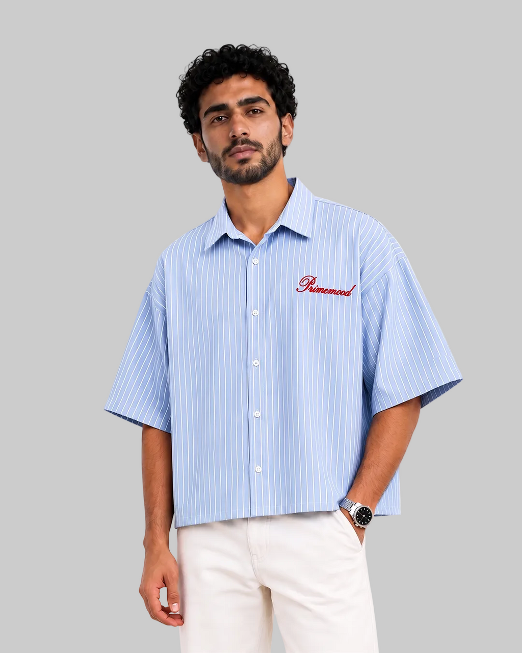 A man wearing a light blue and white vertically striped short-sleeve button-up shirt with 'Primemood' embroidered in red on the chest, paired with off-white trousers, against a plain white background.