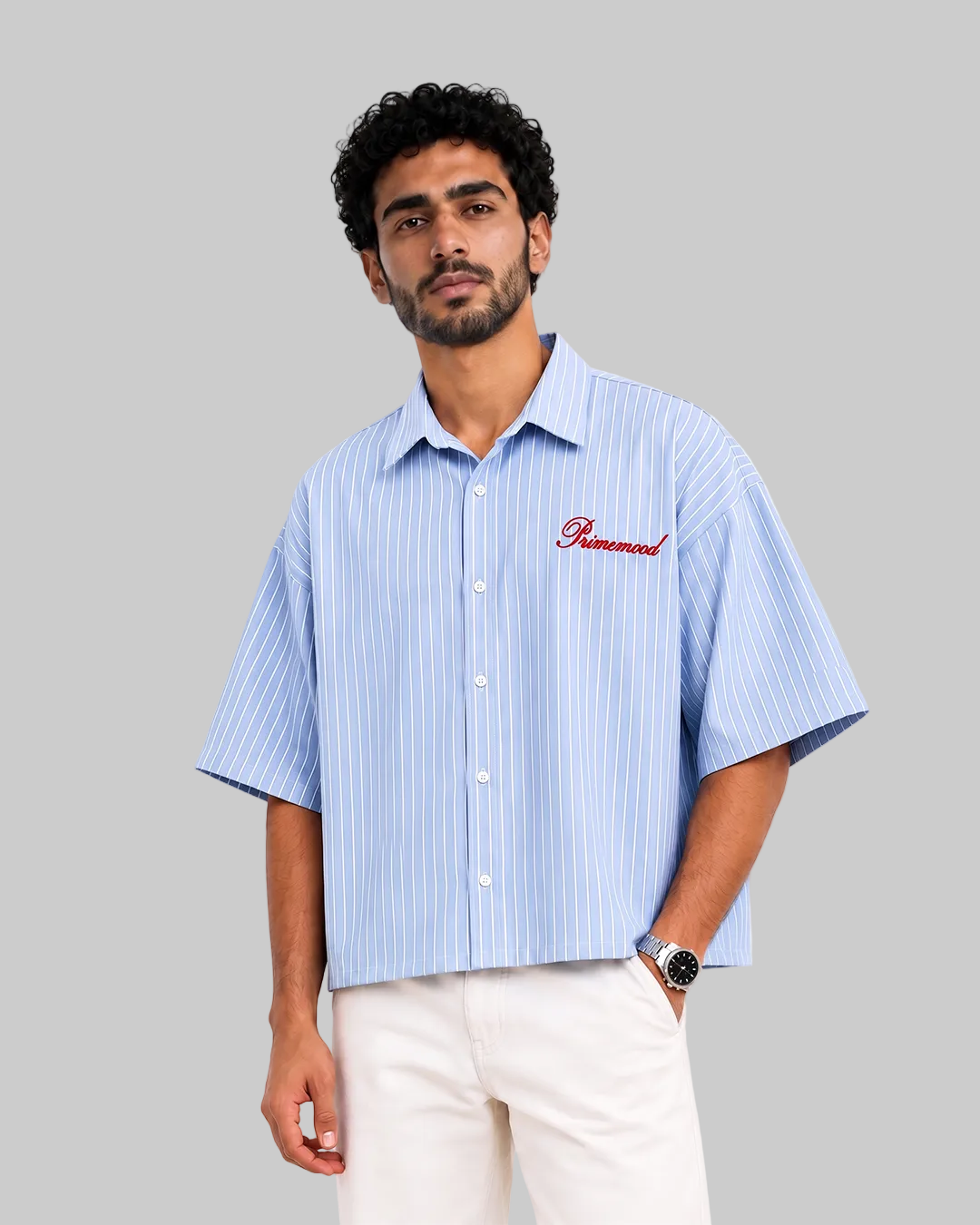 A man wearing a light blue and white vertically striped short-sleeve button-up shirt with 'Primemood' embroidered in red on the chest, paired with off-white trousers, against a plain white background.